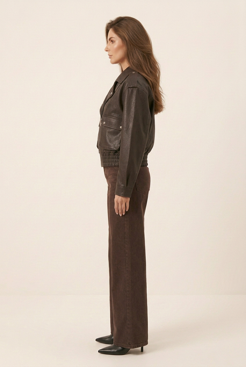 Full-length side profile with the biker jacket and long chocolate-brown jeans draped over black pointed-toe boots